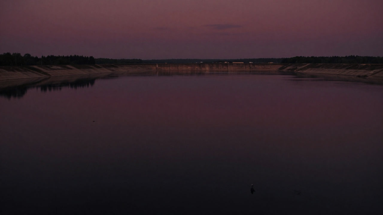 Aerial photo of a desolate dam reservoir. The water surface is calm, yet one can almost hear the ecosystem slumbering beneath.