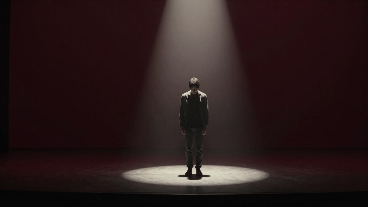 Silhouette of a person standing alone on a dark stage under a single spotlight, looking anxious downward.
